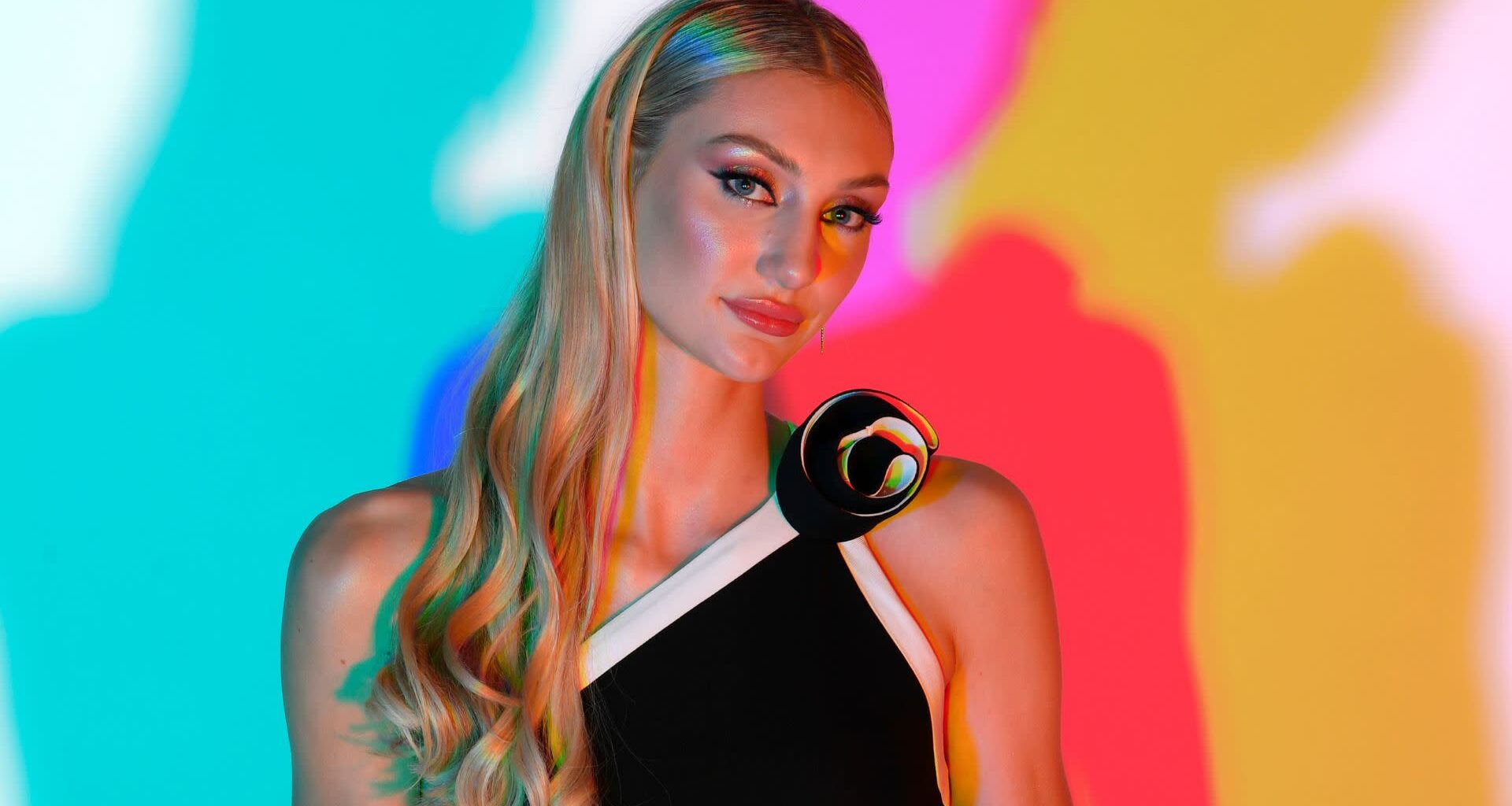 Cameron Brink with long blonde hair in a black and white dress stands against a colorful, abstract background.