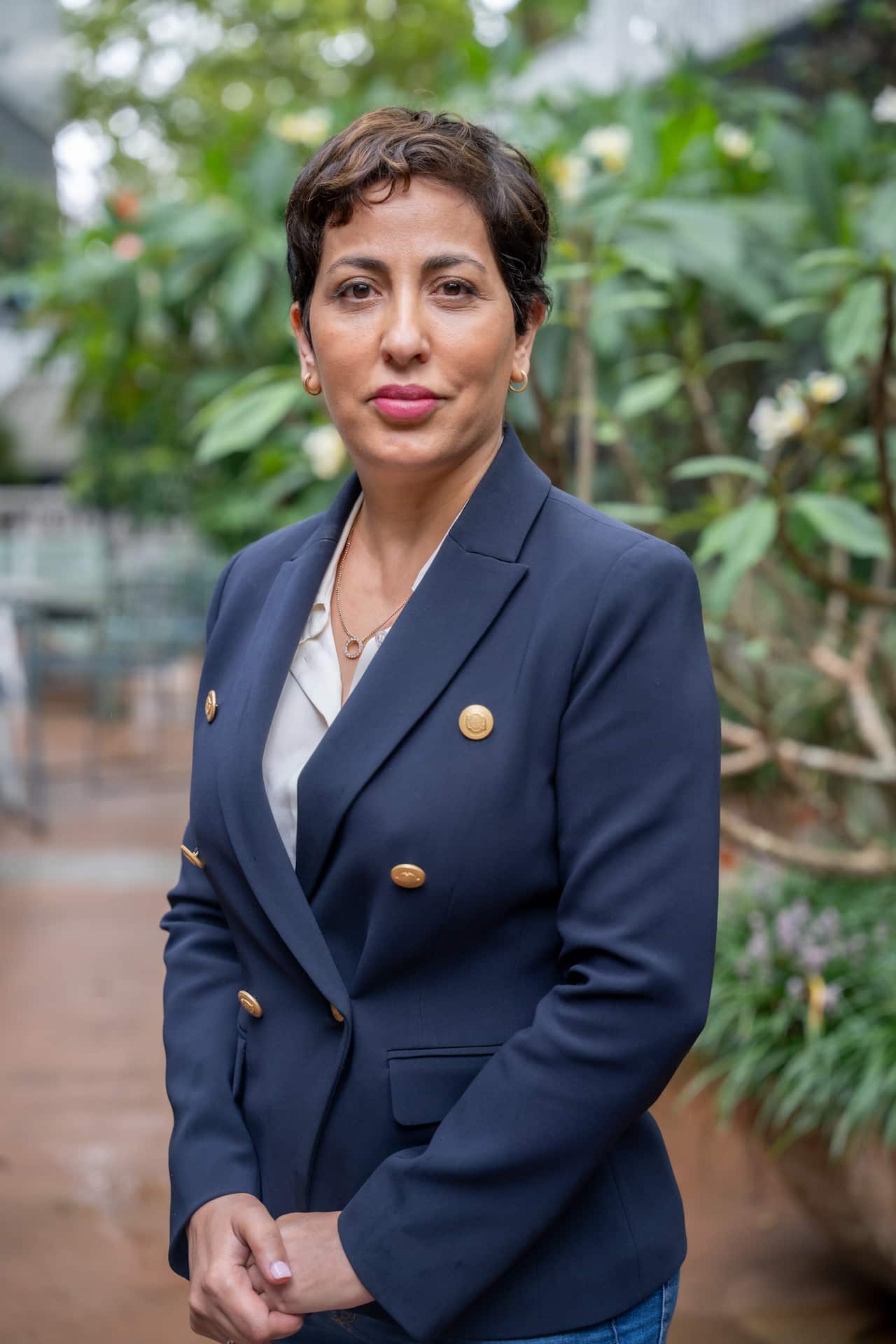 A woman with short brown hair wearing a navy blazer stands in a garden.