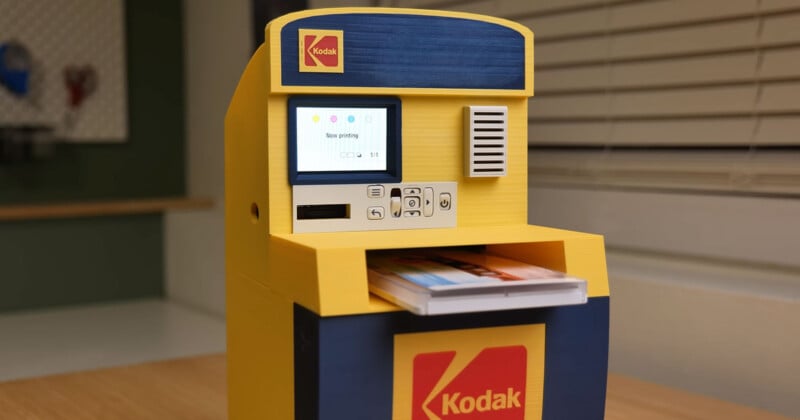 A small yellow and blue Kodak photo printing kiosk sits on a table with printed photos partially ejected from the front slot and a display screen showing a "Now printing" message.
