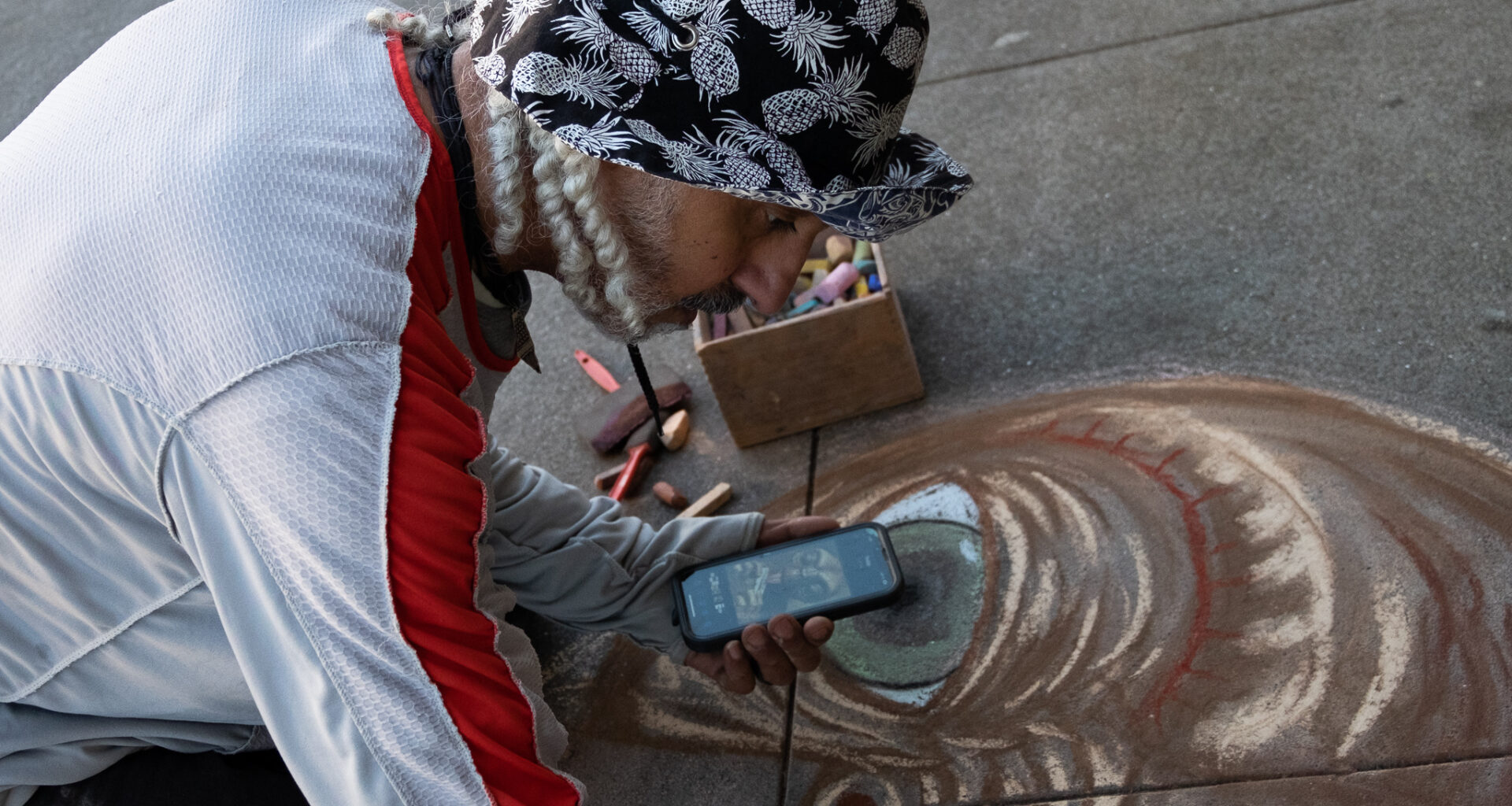 A person wearing a patterned hat and holding a phone creates chalk art on pavement, with pastel sticks and a box nearby.