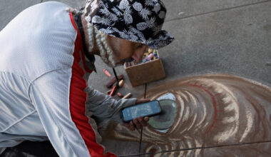 A person wearing a patterned hat and holding a phone creates chalk art on pavement, with pastel sticks and a box nearby.
