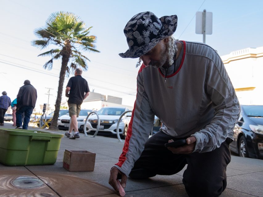A person wearing a patterned hat and long-sleeve shirt kneels on a sidewalk, drawing with chalk. Nearby, a green bin and a wooden box are on the ground. Cars and people are in the background.