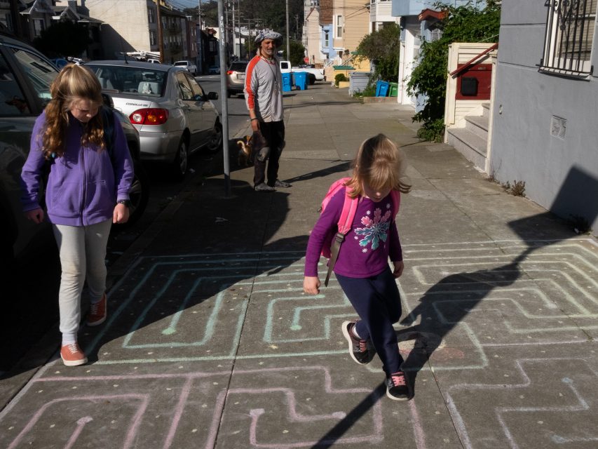 Two young girls walk on a sidewalk decorated with colorful chalk lines, while a man and a dog stand in the background on a sunny street.
