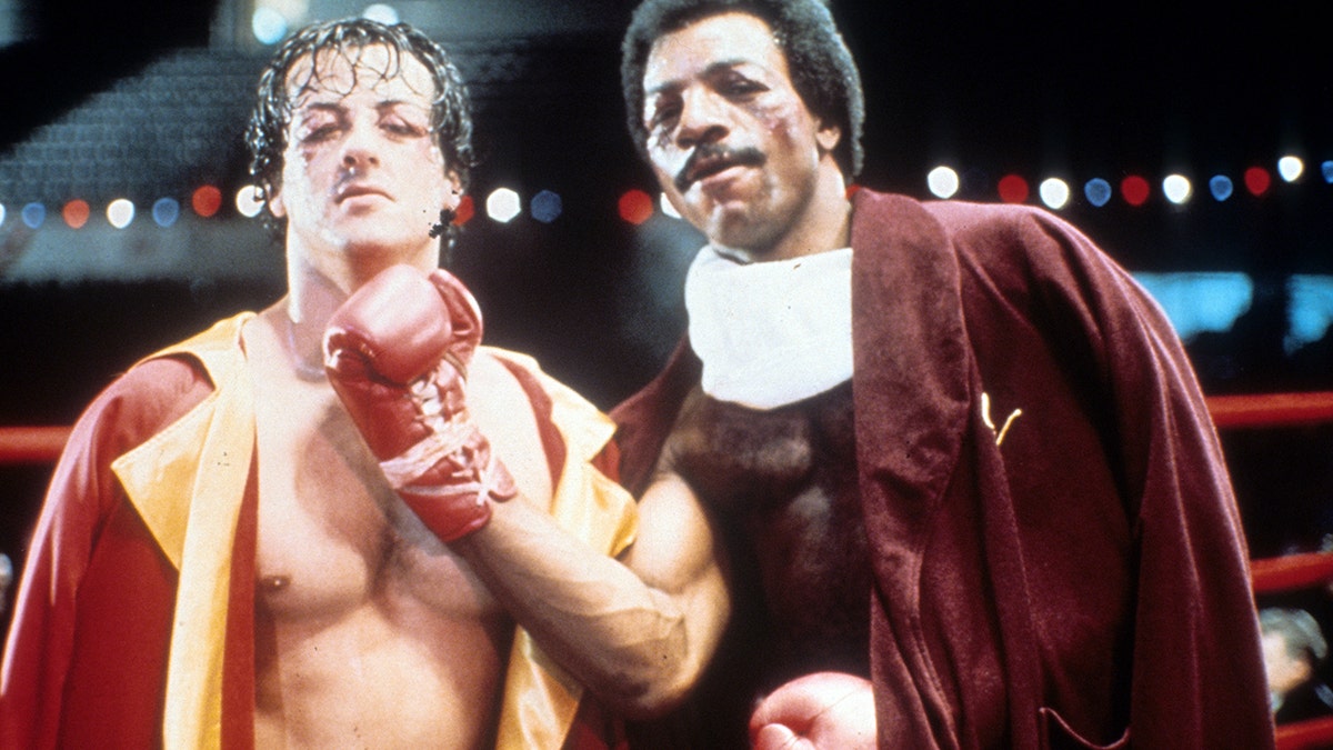 Sylvester Stallone and Carl Weathers, bruised, pose for the camera in the ring