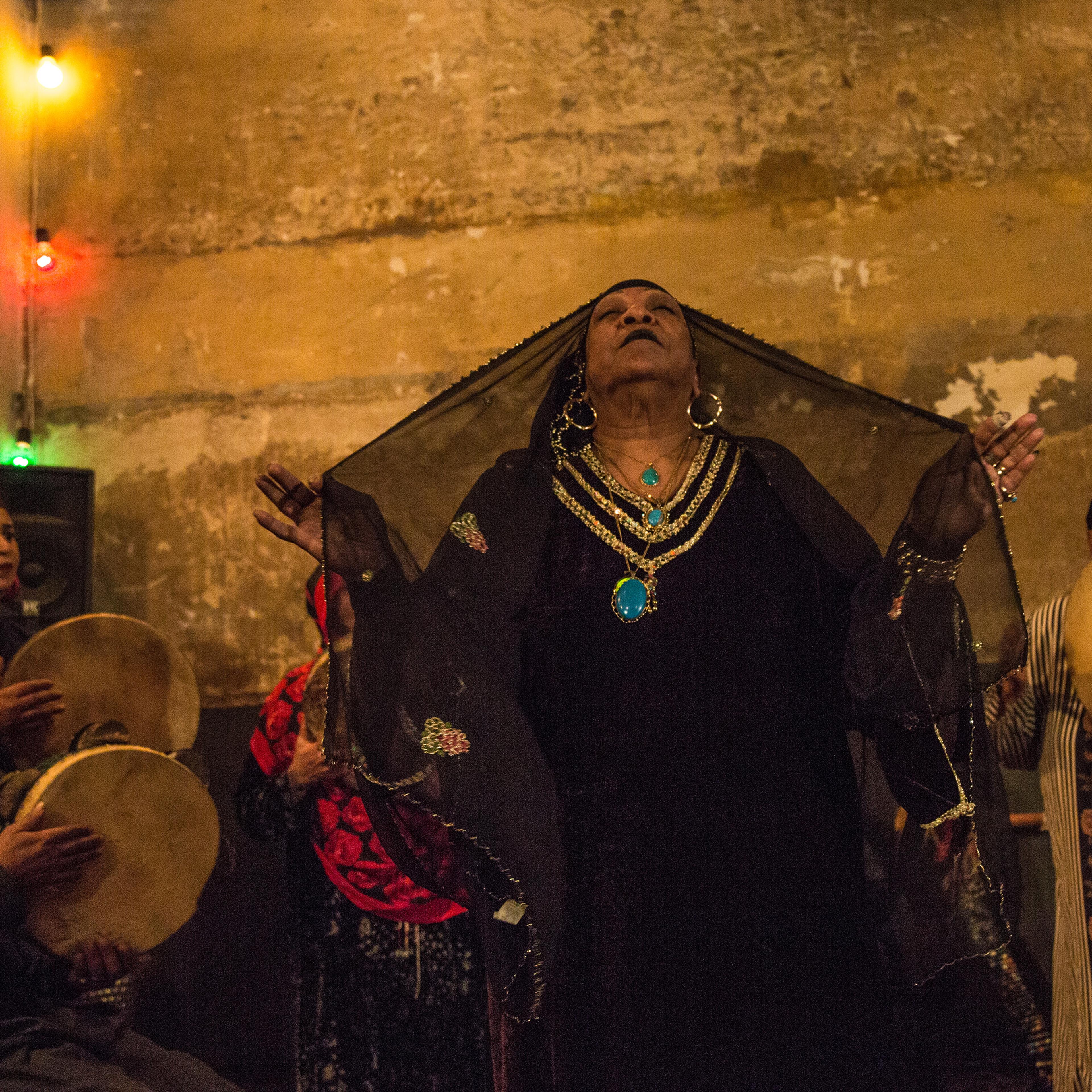 A woman wearing a black gown and veil, and gold and turquoise jewellery raises her harms with eyes closed; musicians wearing headdresses play drums in the background in a dimly lit setting.