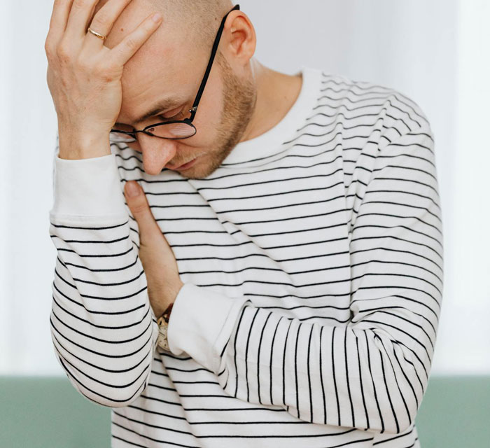 Man wearing glasses and striped shirt looking stressed, holding his head and crossing his arms in an indoor setting. Man wearing glasses and striped shirt looking stressed, holding his head and crossing his arms in an indoor setting.
