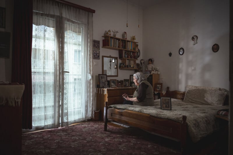 An elderly woman sits alone on a bed in a dimly lit, vintage bedroom, looking at a photo. Family photos and decorations adorn the walls and furniture. Light filters in through lace curtains on a large window.