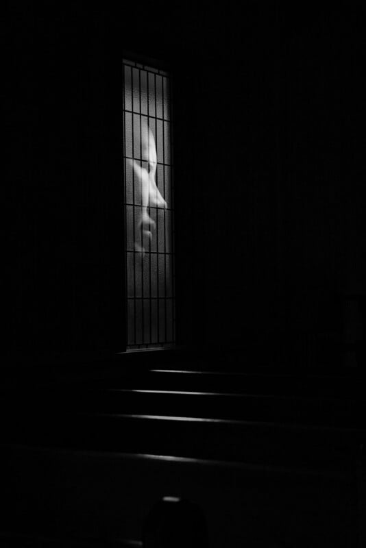 A dimly lit room with empty benches and a single illuminated stained glass window depicting a serene face in profile, creating a calm and contemplative atmosphere.