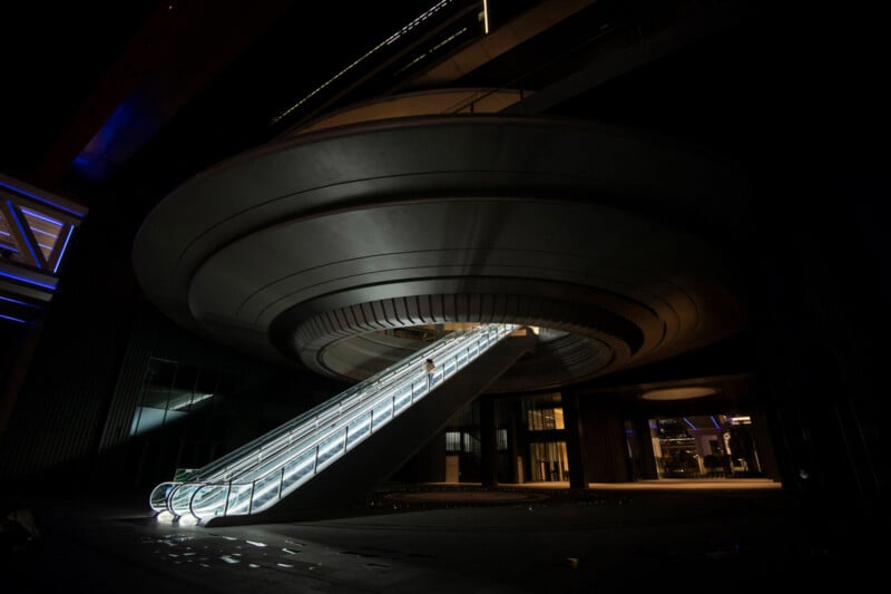 A futuristic, circular structure with a glowing escalator leading up to it, illuminated at night; the architecture resembles a UFO or spaceship.