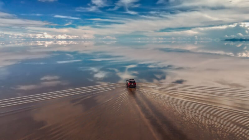 A vehicle drives across a vast, reflective salt flat, creating ripples in shallow water under a blue sky with scattered clouds, blending seamlessly with the horizon.