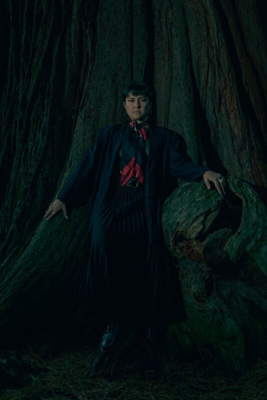 A person in a dark, flowing outfit and red scarf stands against the trunk of a massive tree, with one hand resting on a large twisted root in a dimly lit, forest environment.