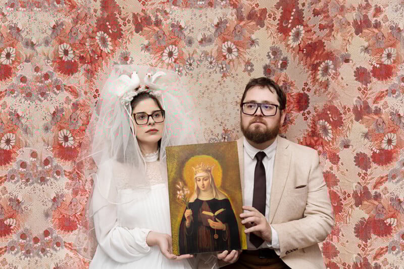 A solemn couple in vintage attire poses against a floral-patterned backdrop; the woman wears a white dress with a veil and decorative doves, while the man wears a light suit and glasses. They hold an icon of a crowned saint.