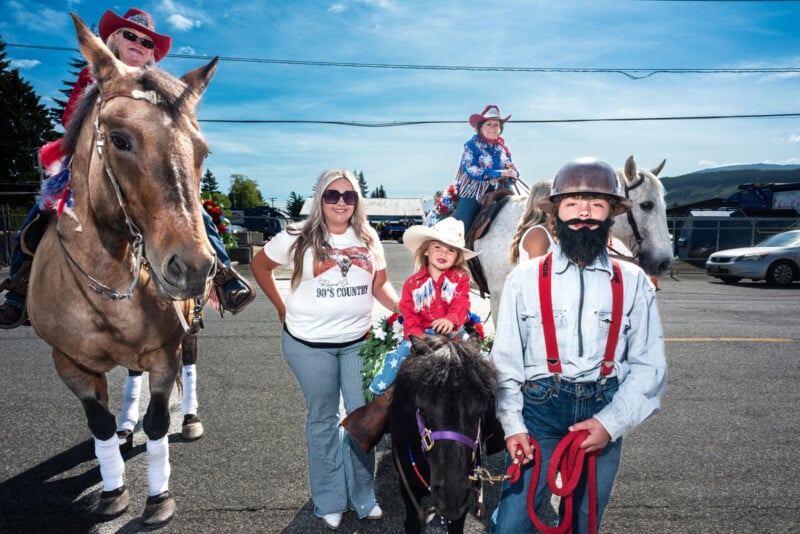 A group at a parade: two adults on horses, a smiling woman, two children dressed in cowboy attire (one with a fake beard) and one child on a small pony, all in western-themed clothing on a sunny day.