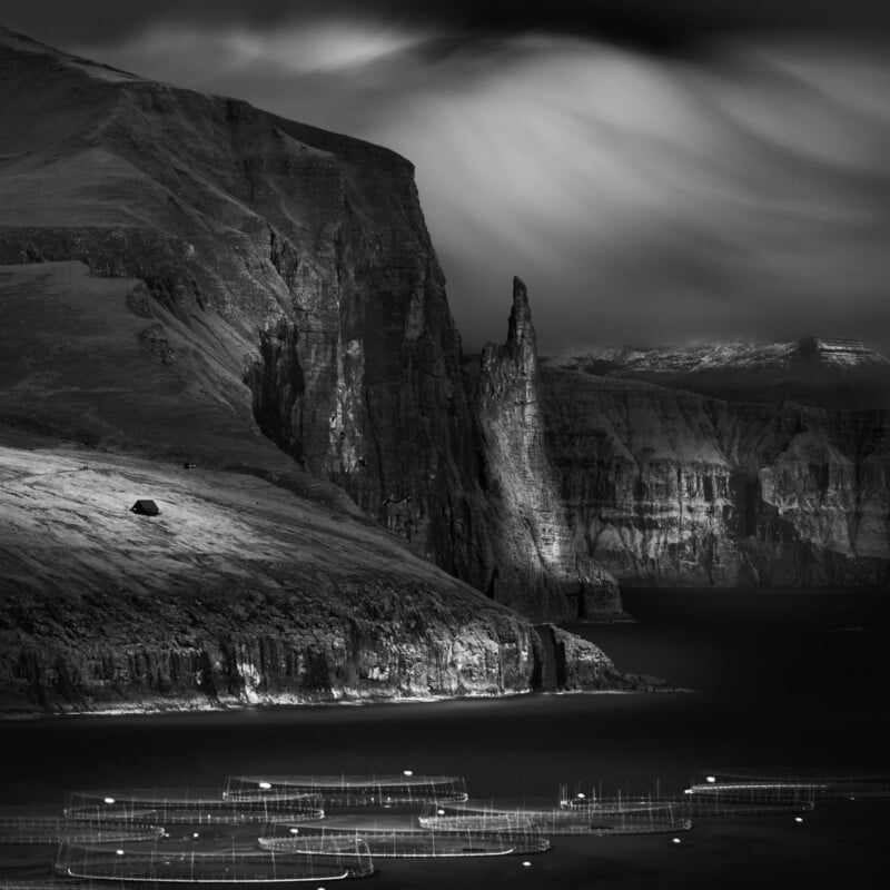 Dramatic black-and-white photo of steep cliffs rising above a calm sea, with vertical rock formations and soft, streaked light from above. Fish farm nets float in the water in the foreground.