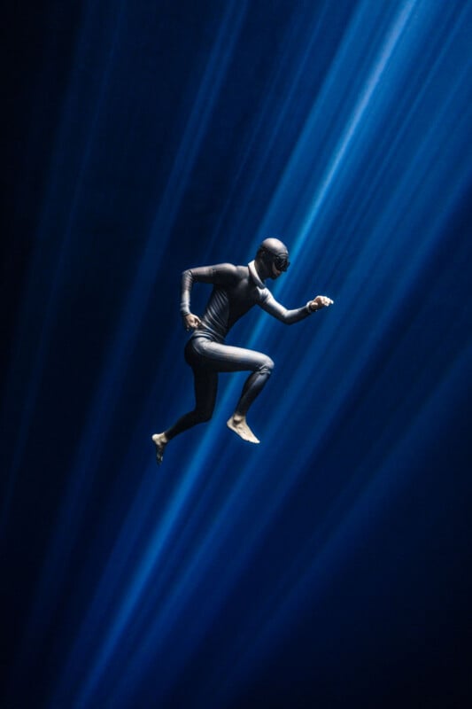 A person in a black wetsuit, mask, and fins is suspended underwater, appearing to run through beams of blue light that shine diagonally from above. The deep water surrounds them in a dramatic, surreal scene.