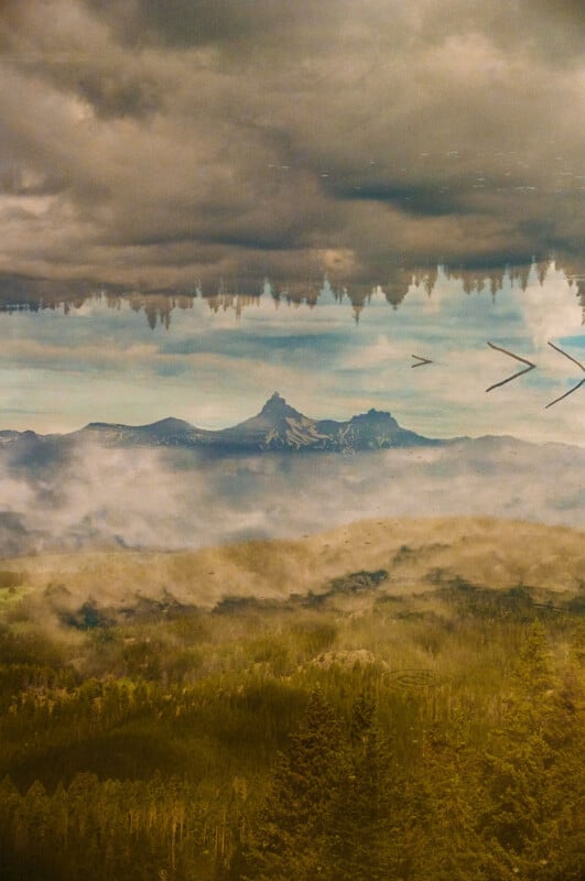 A misty mountain landscape with a forest in the foreground and dramatic clouds above, reflected in a calm body of water. The reflection creates an almost surreal, upside-down effect.