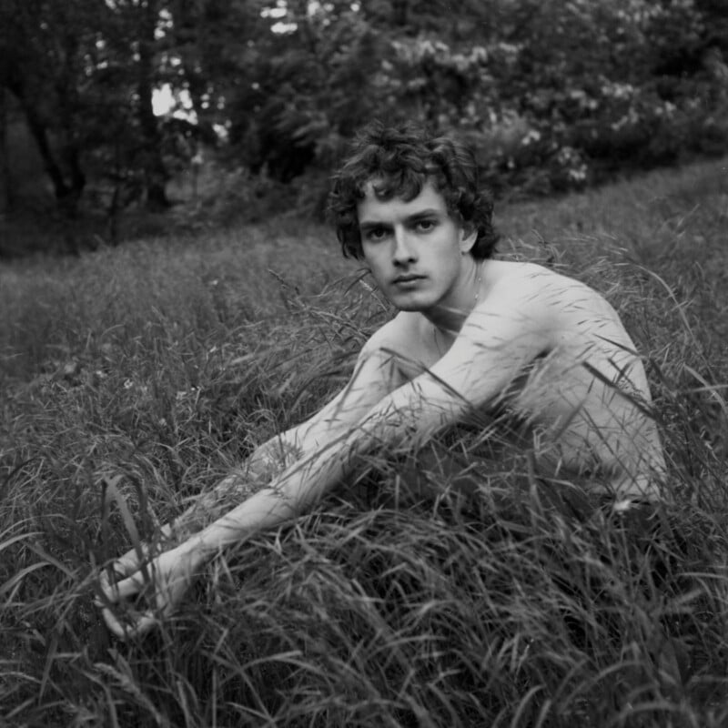 A young man with curly hair sits shirtless in tall grass, looking directly at the camera. Trees and foliage form a blurred background in this black and white photo.