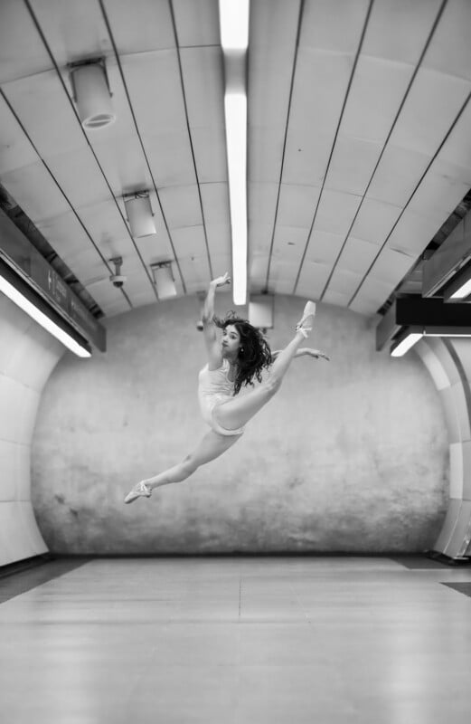 A ballerina in a leotard gracefully leaps mid-air in an arched, modern subway tunnel. The photo is in black and white and captures her poised form, outstretched arms, and pointed toes beneath bright ceiling lights.