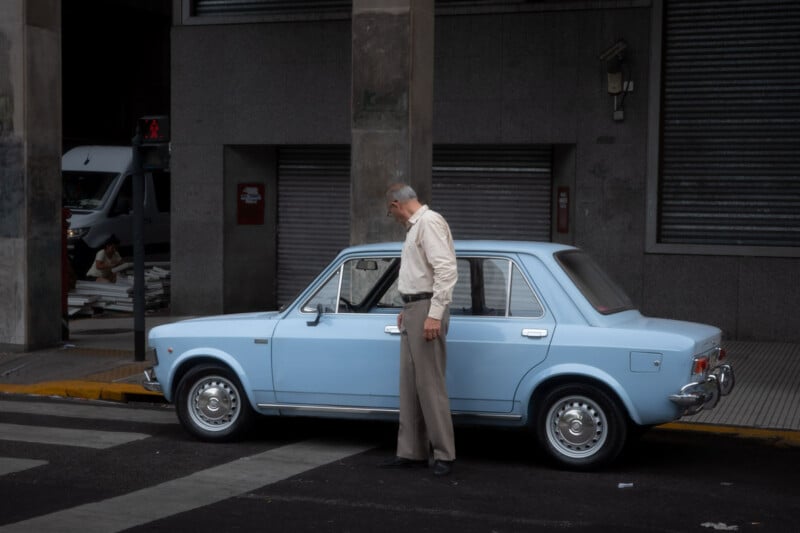 An older man in a light shirt and beige pants stands beside a light blue vintage car parked at a curb in an urban area with gray buildings and closed metal shutters in the background.