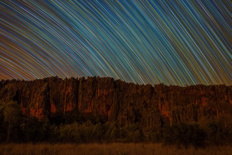 Colorful star trails streak across a night sky above a rugged, rocky cliff and sparse vegetation, creating an arc pattern and highlighting the dramatic landscape below.