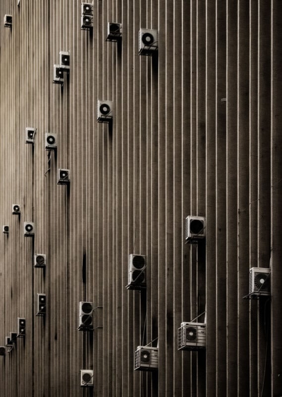 Rows of air conditioning units are mounted on the exterior of a tall, vertically-striped building wall, creating a geometric and repetitive pattern in a sepia-toned image.