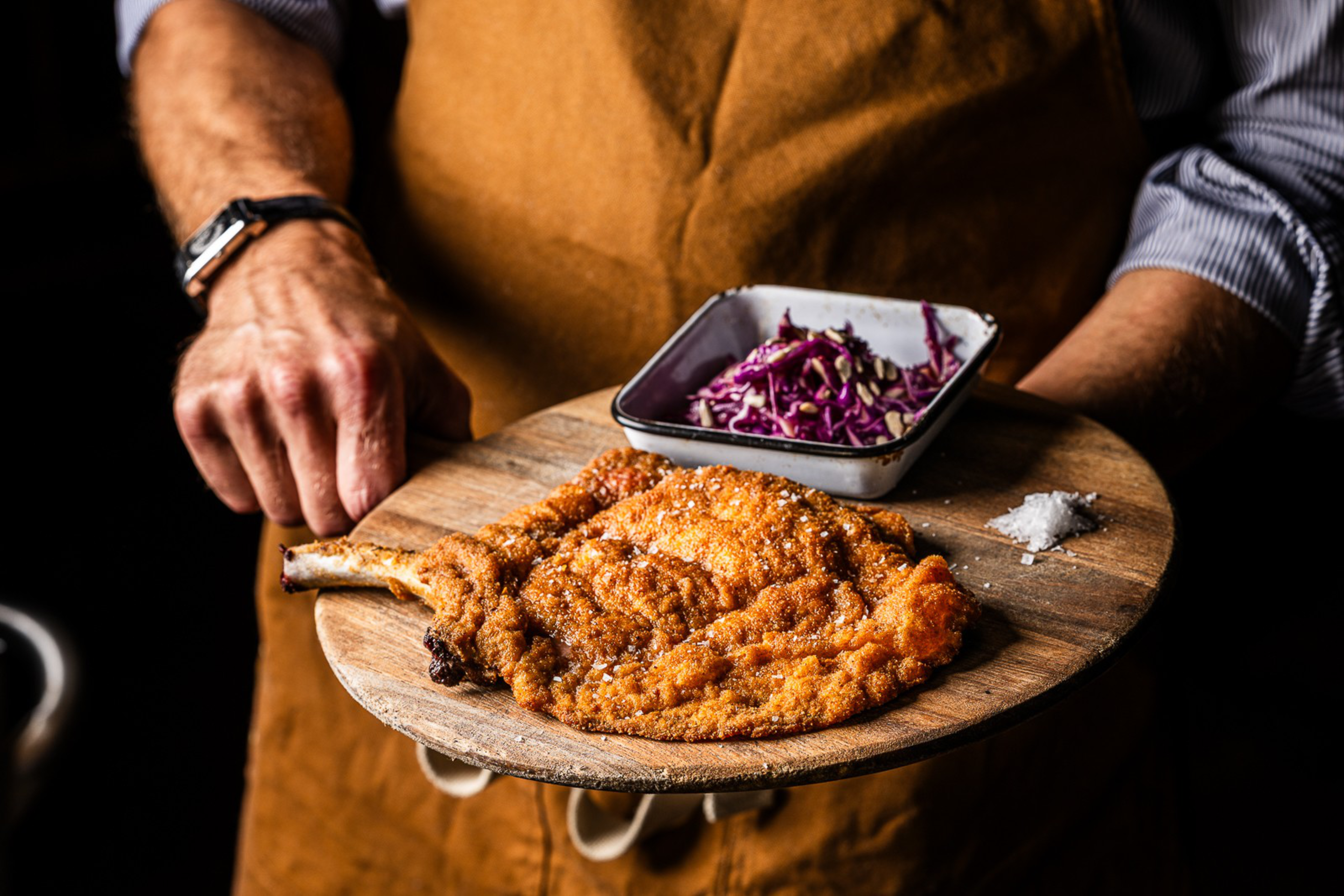 A person wearing a brown apron holds a wooden board with a large breaded pork cutlet, a small bowl of purple cabbage slaw, and a small pile of coarse salt.