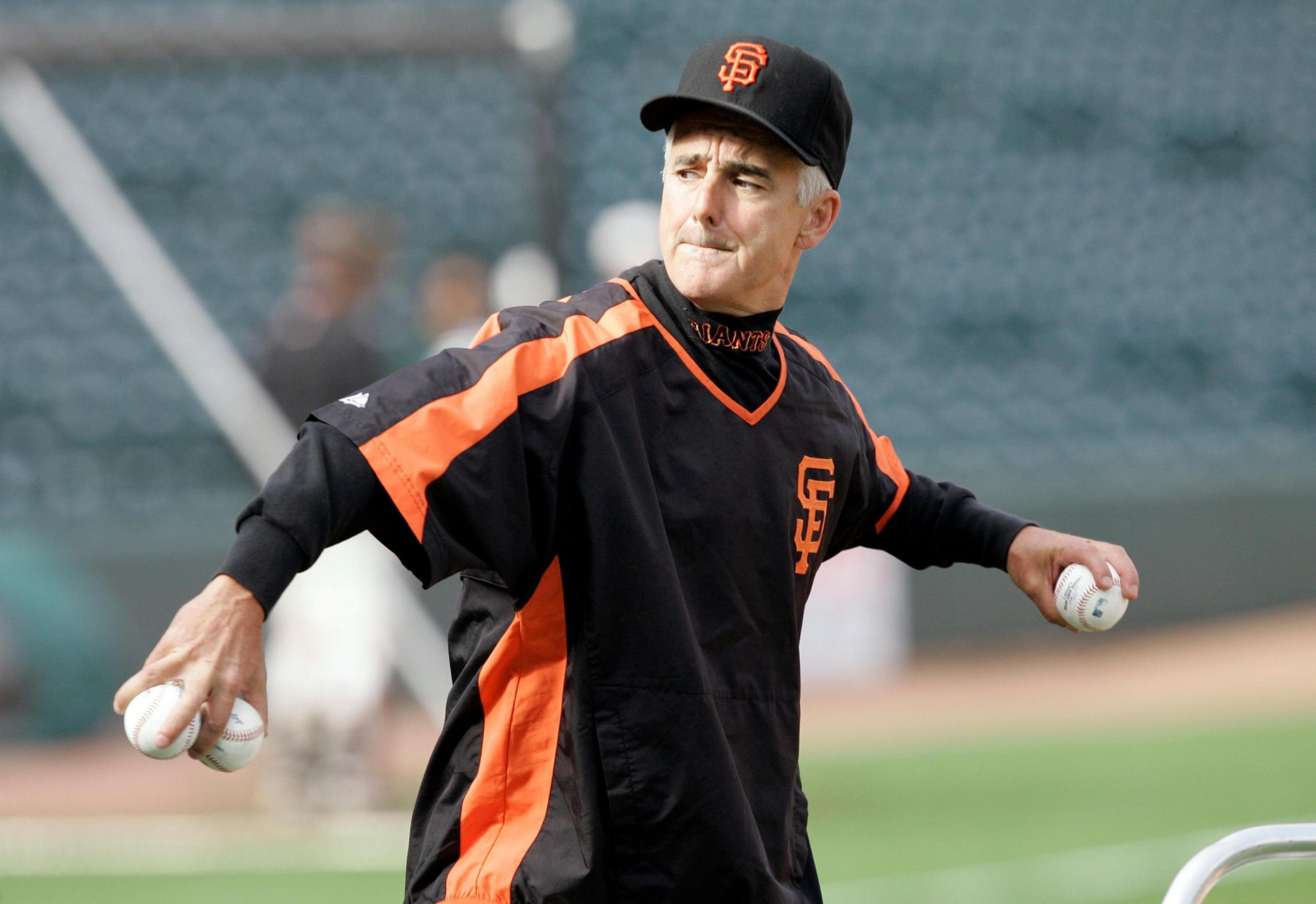 A man in a San Francisco Giants black and orange jacket and cap holds three baseballs, preparing to throw one on a sports field.