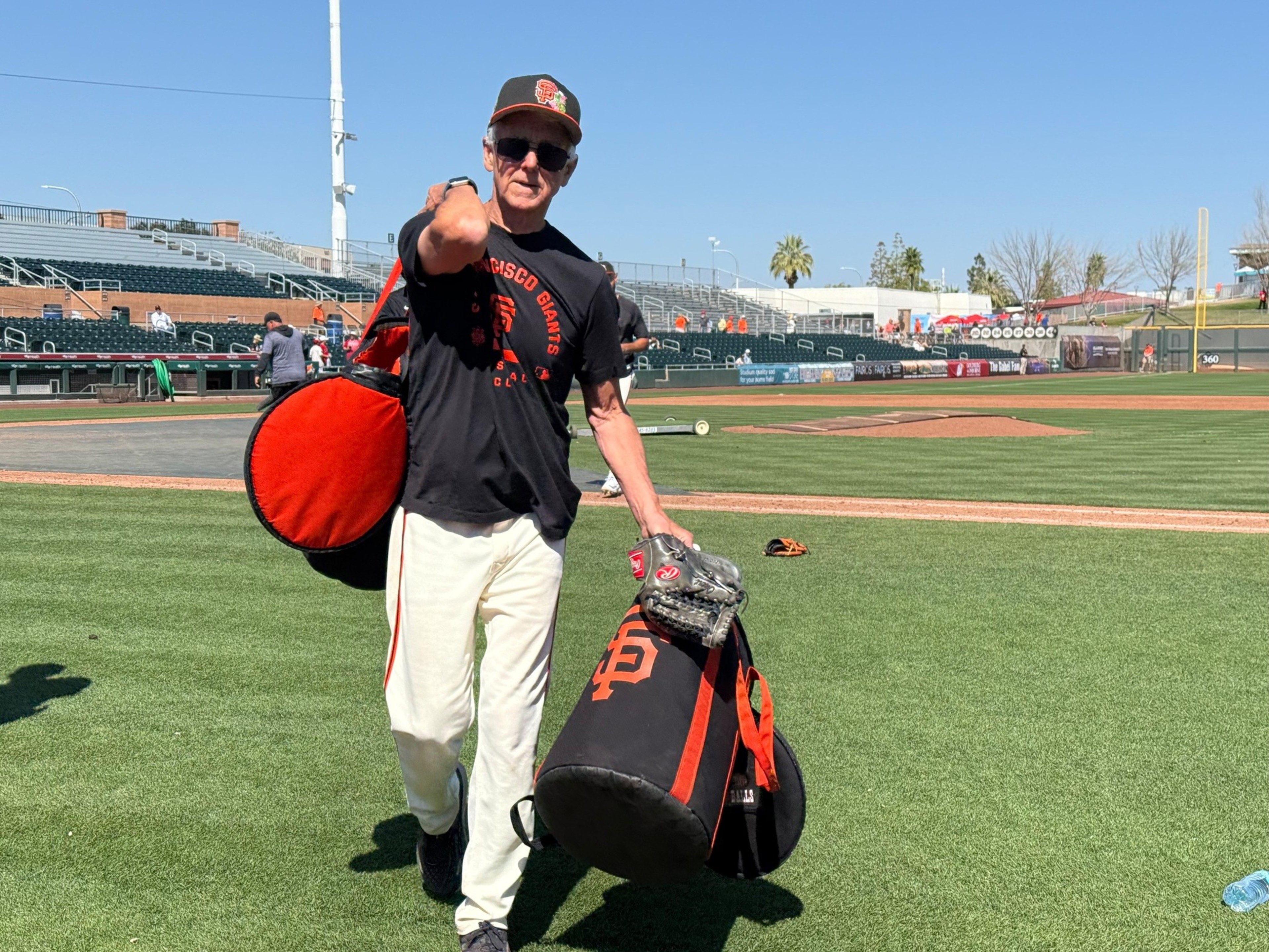 An older man wearing a San Francisco Giants cap and shirt carries two large bags and a baseball glove on a sunny baseball field.
