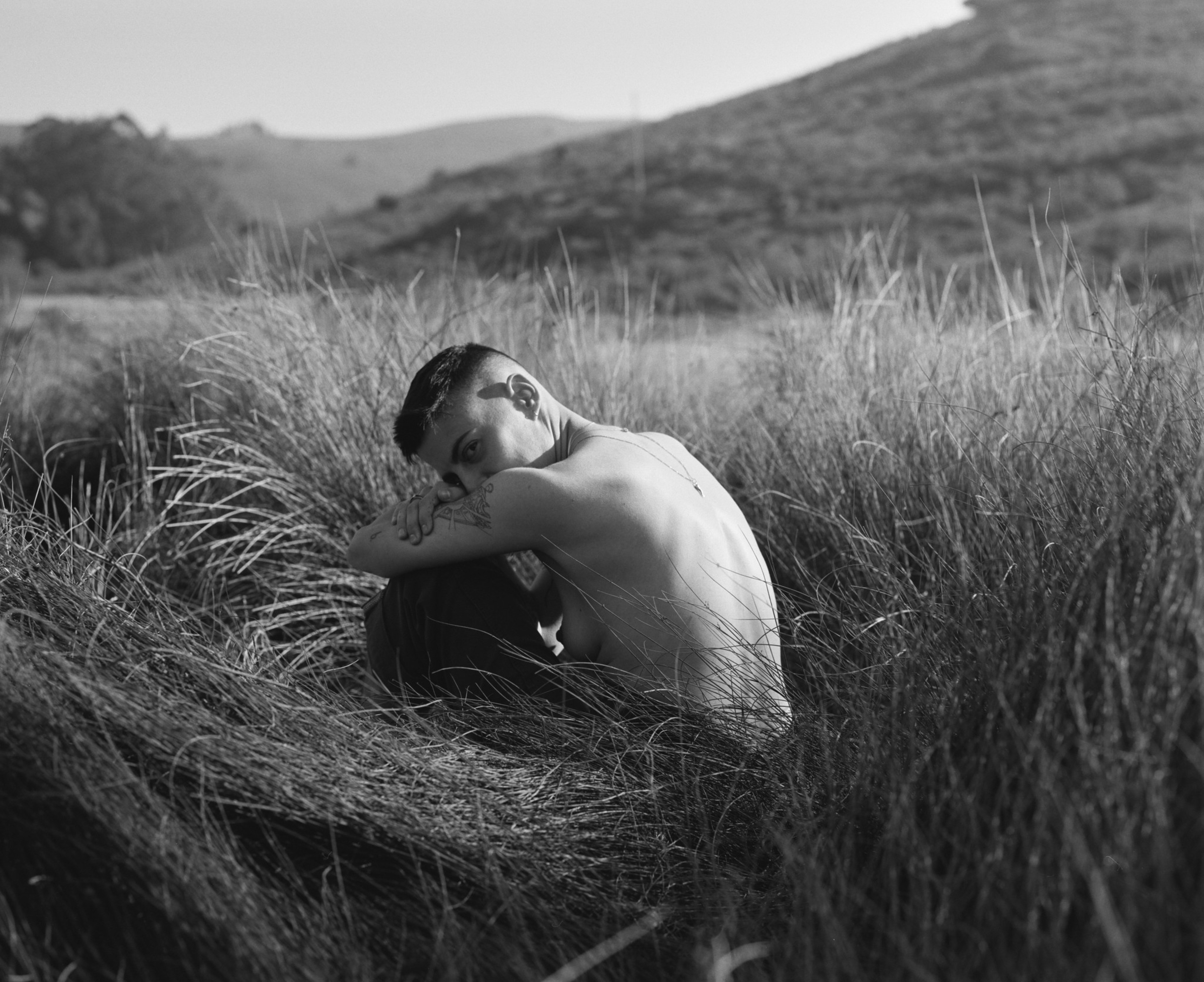 A shirtless person with short hair sits curled up in tall grass, resting their head on their arm while looking back over their shoulder.