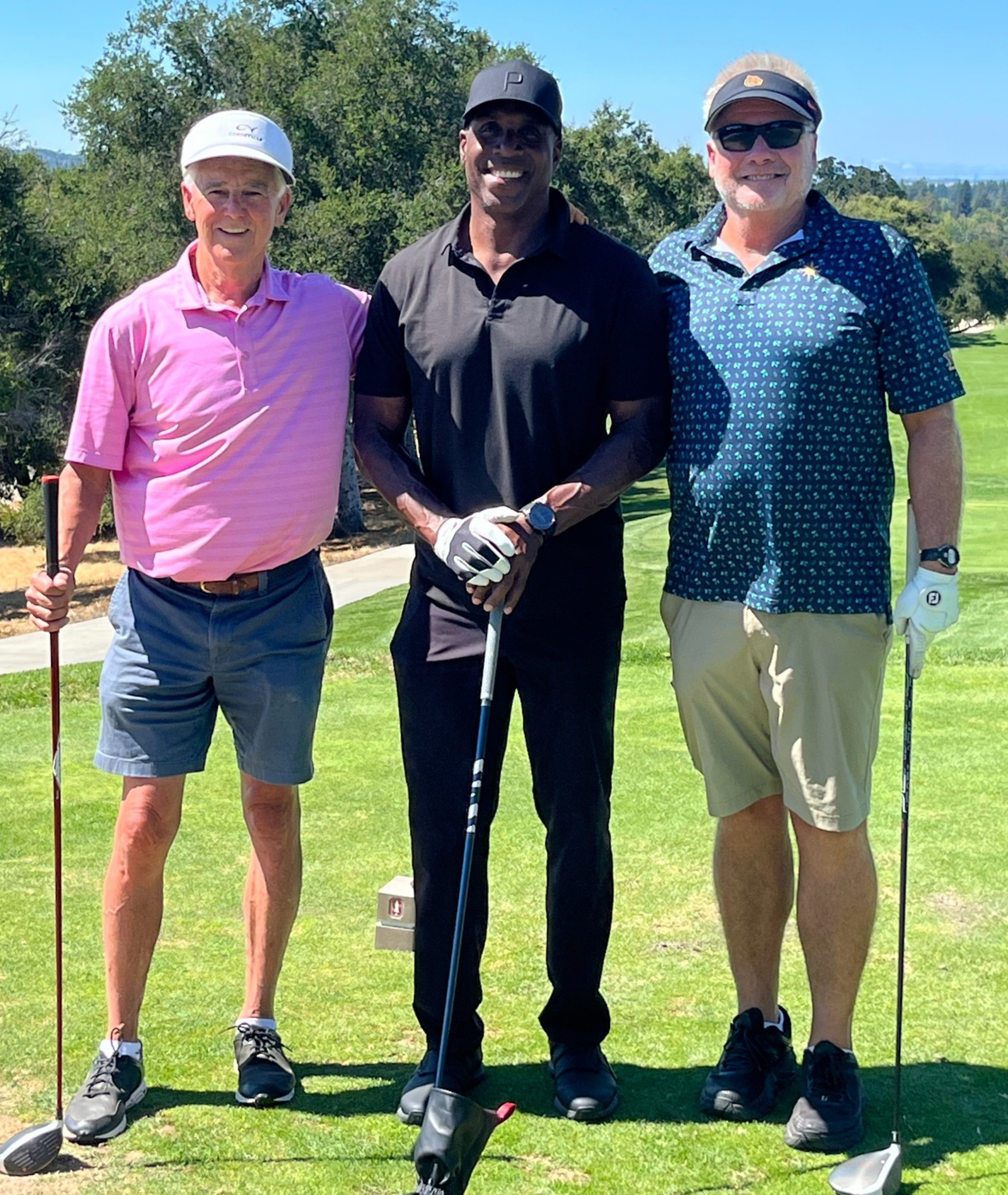 Three men stand on a golf course holding golf clubs, dressed in polo shirts, shorts or pants, with trees and a blue sky in the background.