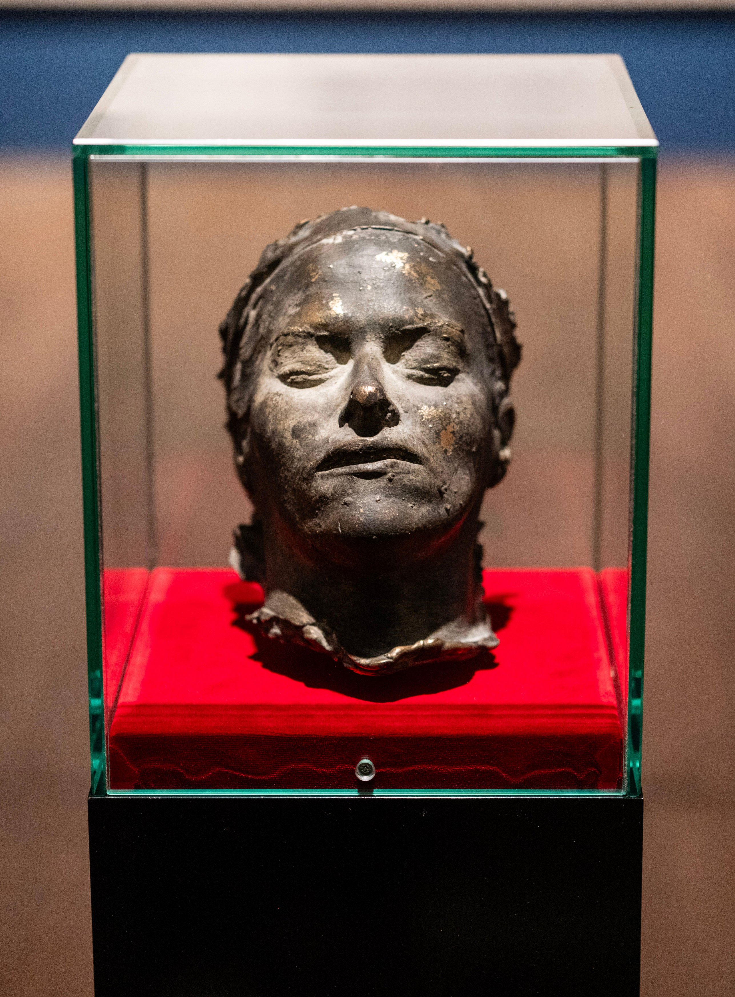 A bronze death mask with closed eyes and a somber expression displayed in a glass case with a red velvet base.
