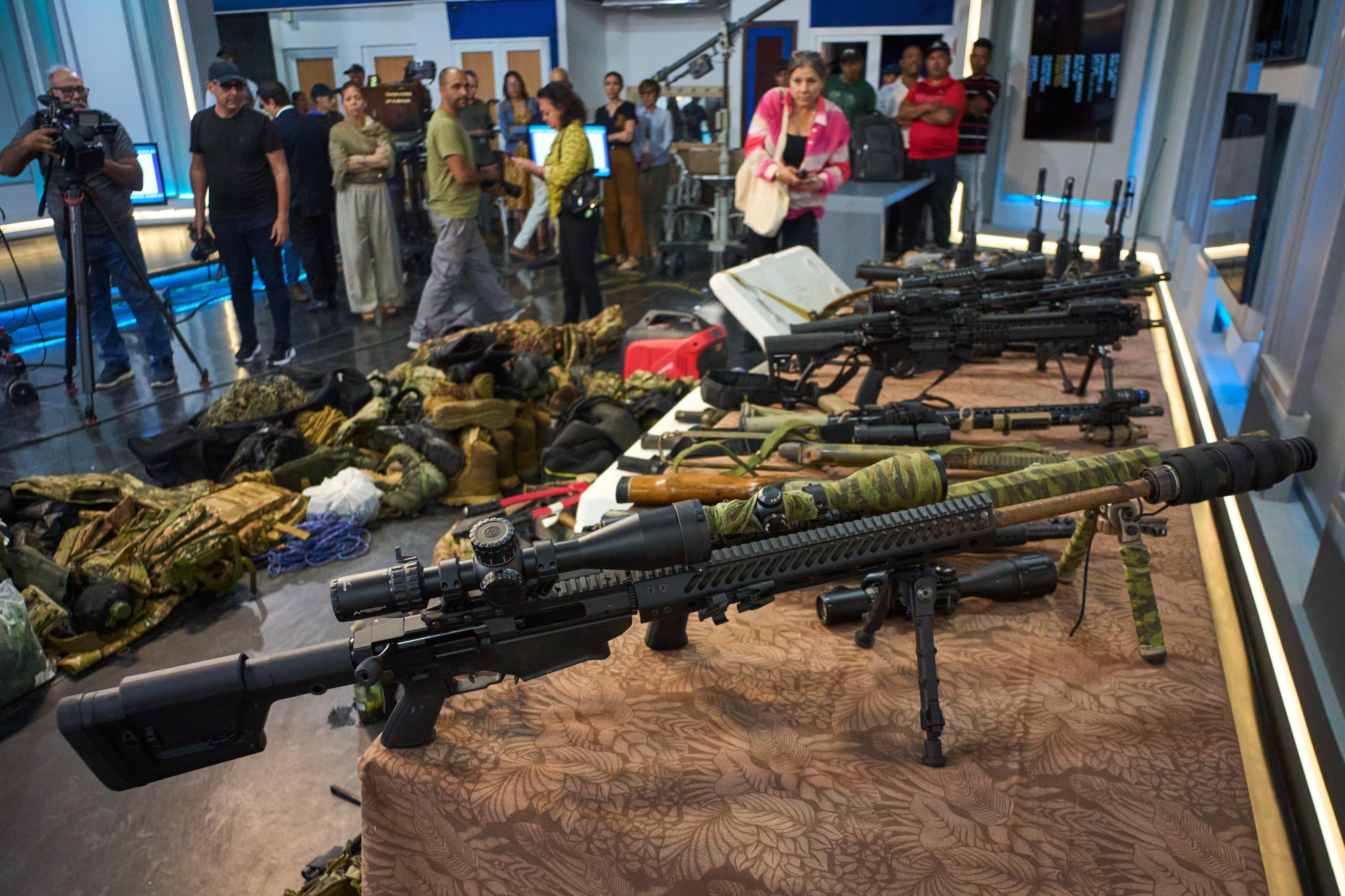 Assorted weapons, including rifles and camouflaged gear, displayed on a table with a patterned cloth, while people gather in the background.