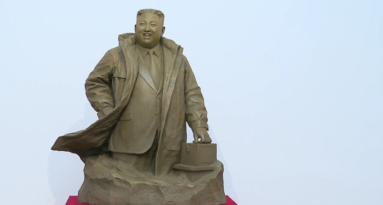 Sculpture of Kim Jong Un in a trench coat, with one hand in his pocket and the other resting on a small box.