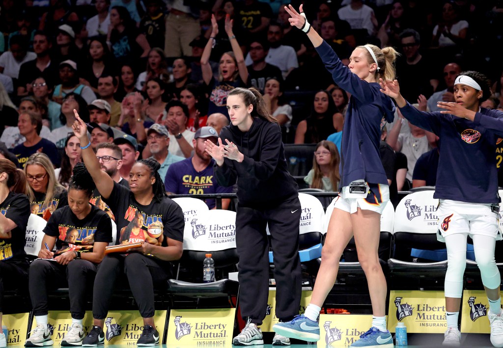 Caitlin Clark, in plain clothes, on the bench with other Indiana Fever players and staff, watching the game and cheering.