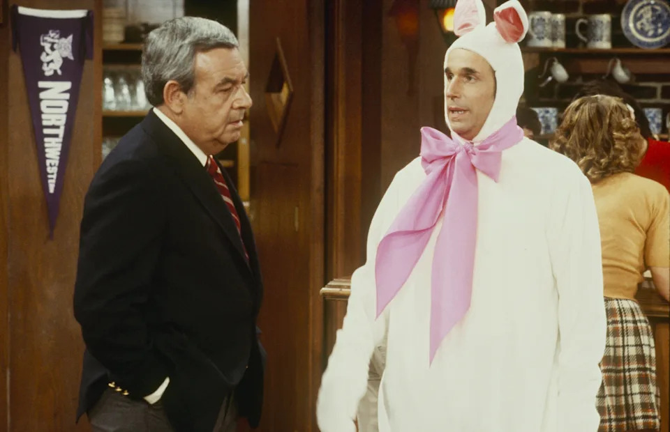A man dressed in a bunny costume with a pink bow talks to another man in a suit inside a room with a wooden interior