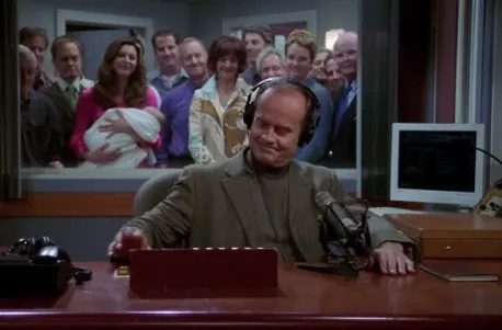 A man wearing headphones, seated at a radio station console, smiles as a group of people, including someone holding a baby, watches through a window