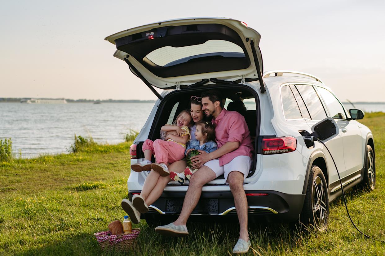 Happy family on the summer beach holiday sitting in the car trunk, waiting for electric car to charge at the vacation spot.