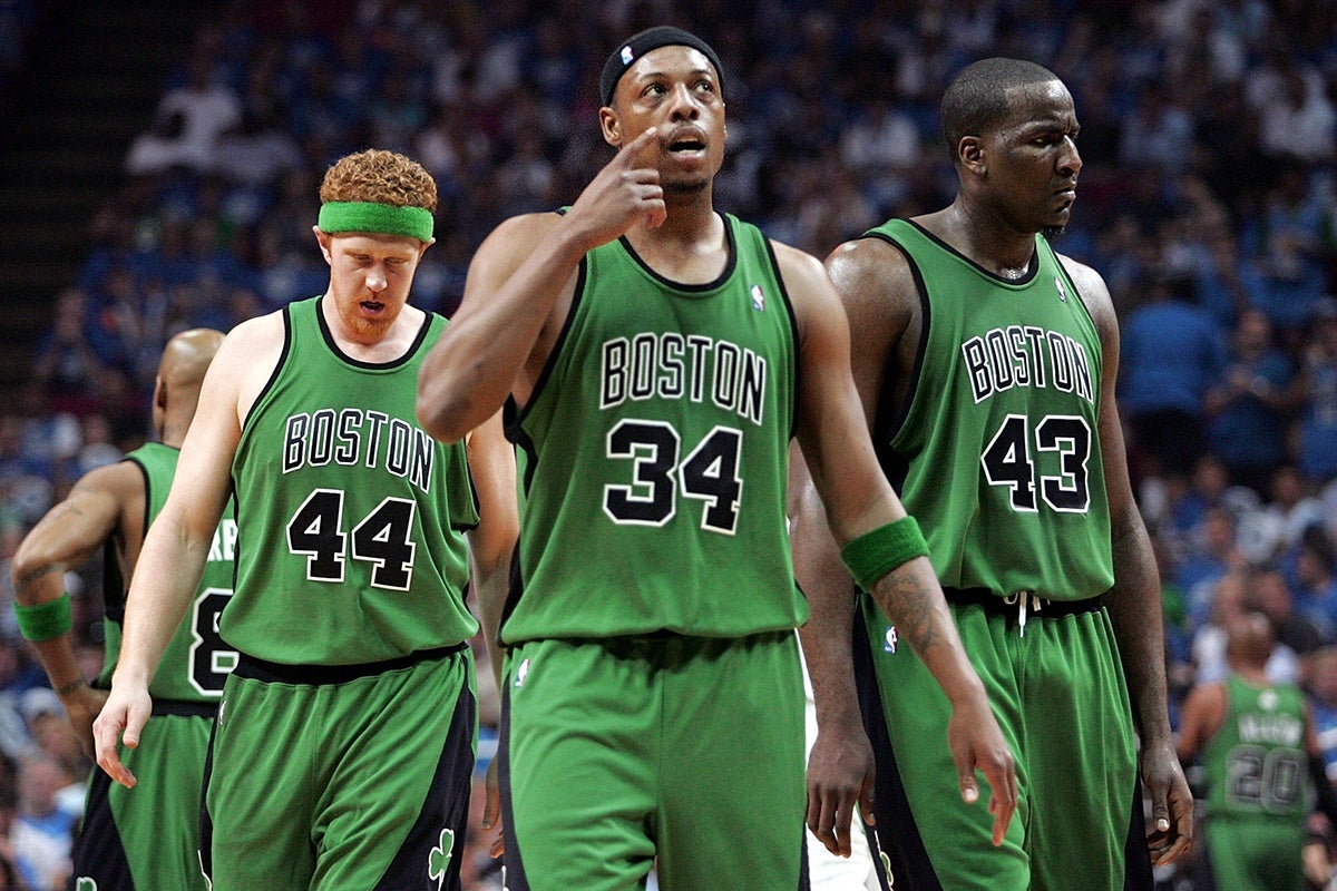 Three Boston Celtics basketball players in green uniforms are on the court during a game. The player in the center, wearing number 34, is pointing upward with his right hand. To his left is a player with red hair and a green headband, wearing number 44, looking downward. To the right is a player wearing number 43, looking to the side. The background shows a blurred crowd in the arena.
