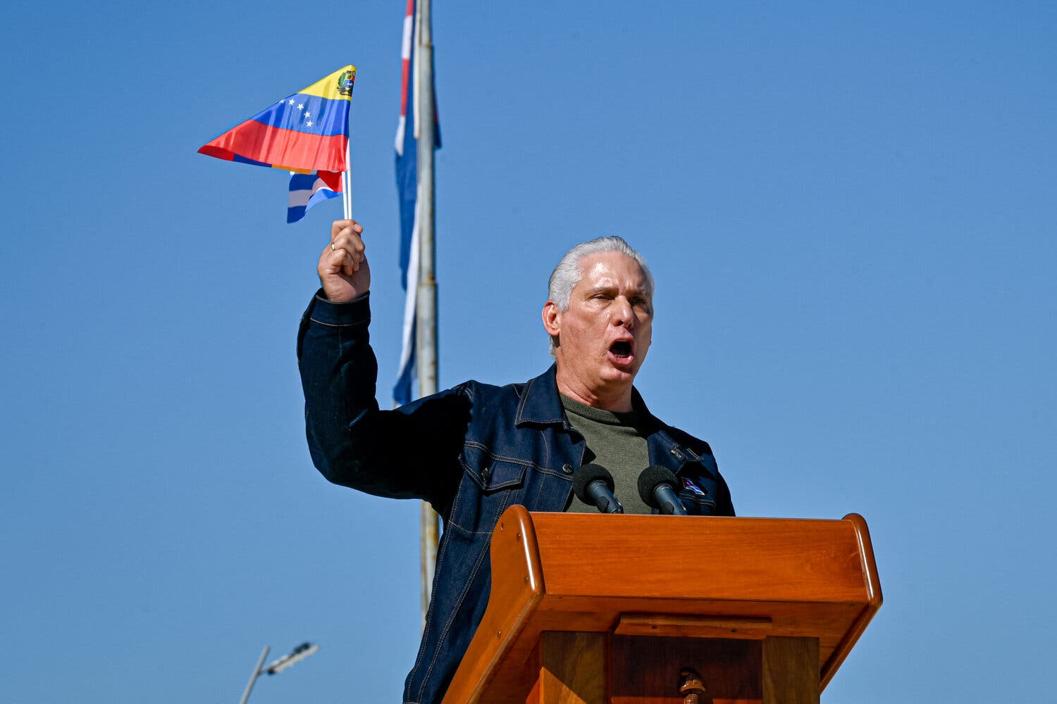 A person in a denim jacket stands at a podium, speaking with an open mouth. They raise a hand holding a small, colorful flag.