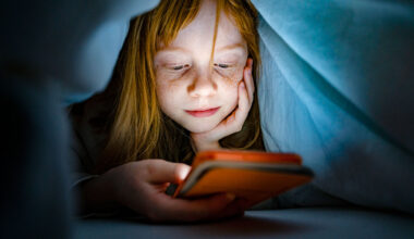 a pale child with freckles hides under a blanket to use a cell phone at night