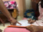 A young girl writes in a notebook as a pointed finger directs her to a line on the page.