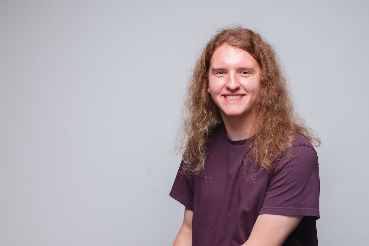 Head and shoulders portrait of a young, Caucasian woman with long, curly blond hair. She is wearing a plum-coloured T-shirt. 