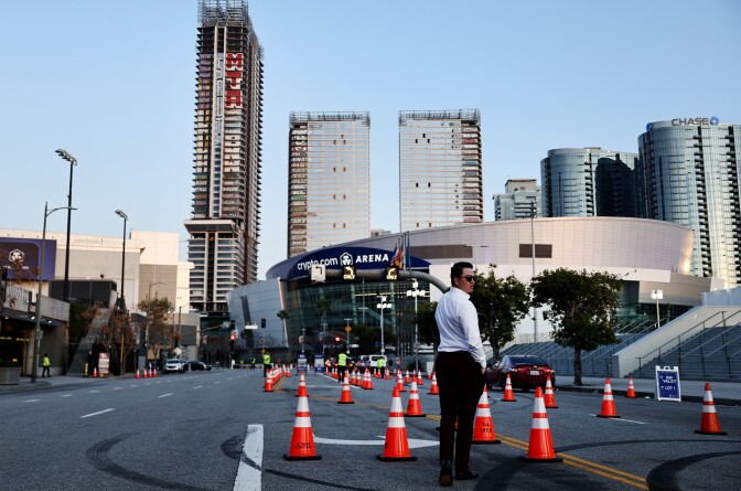 Man in white shirt and black pants walks through road closed off by orange cones near Crypto.com Arena in Los Angeles. In the background are three unfinished skyscrapers.
