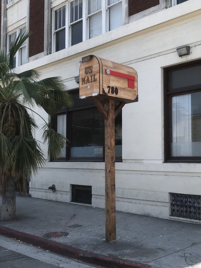 An oversized wooden mailbox sculpture labeled “U.S. Mail” stands on a tall post along a sidewalk.