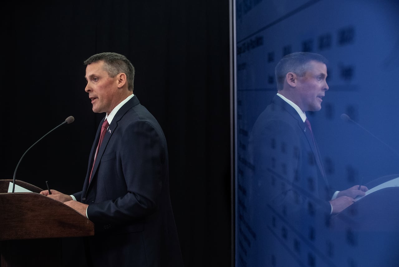 Person speaks at lectern with his reflection in a screen filled with numbers