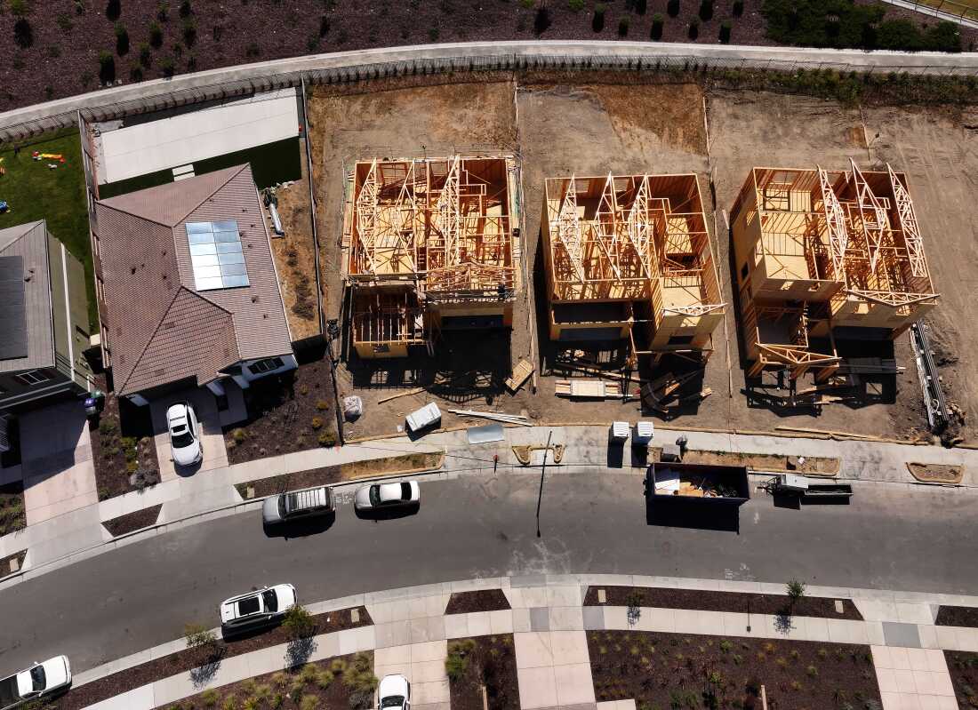 This aerial-view photo shows three homes under construction. The homes are made of wood framing and don't yet have roofs. A complete home is on the left side of the frame.