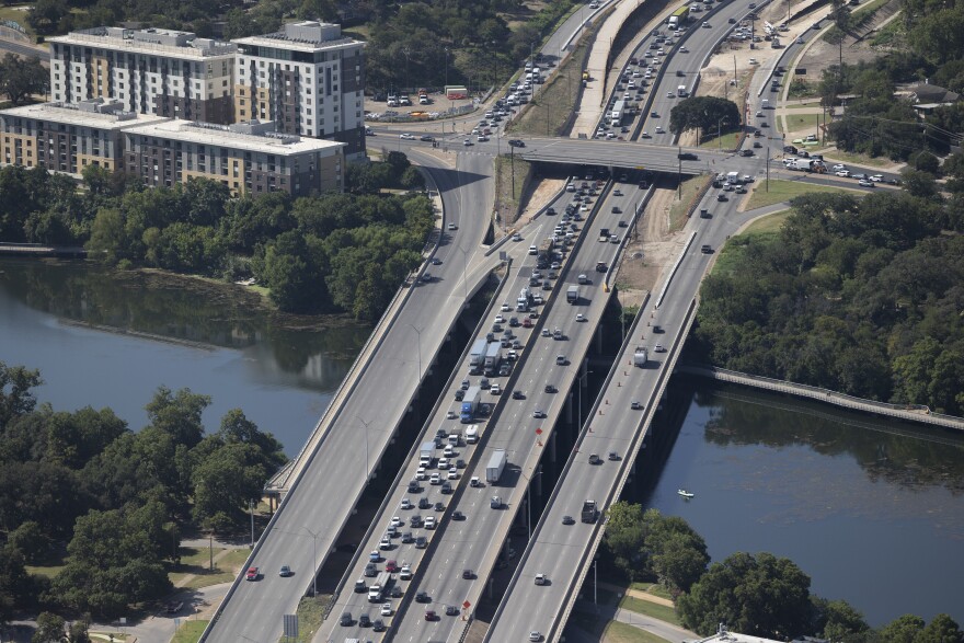 A view of the I-35 bridge over Lady Bird Lake from the top floor of the Waterline building in July 2025. It's daytime. Northbound traffic is congested. Southbound traffic is free flowing. Lady Bird Lake, lined by trees, is reflecting tints of dark green. 