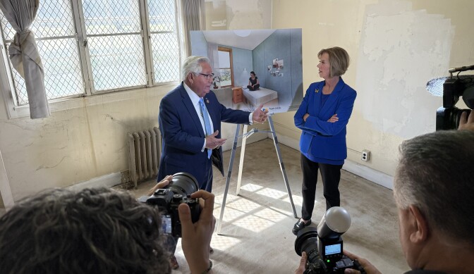 A man in a blue suit stands beside a woman wearing a bright blue jacket. They stand in an empty room and before a poster of furnished room. 