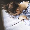 A student appears fatigued while studying late at night, resting her head on a textbook with notes scattered around, reflecting the pressure of academic demands.