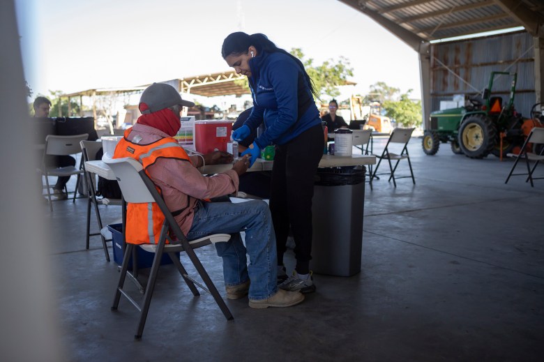A medical personnel member places a blood sugar test on a person's hand while the person faces away. In the background, a mobile medical unit is inside an equipment barn on a farm.